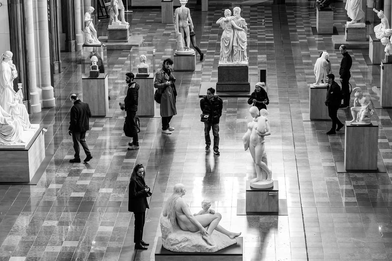 Galeria de museu com esculturas clássicas e visitantes, em preto e branco.